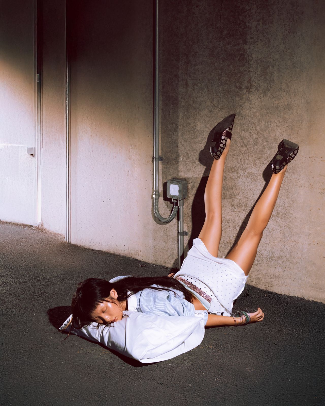 A woman asleep on a pillow on the asphalt, legs raised against a concrete wall